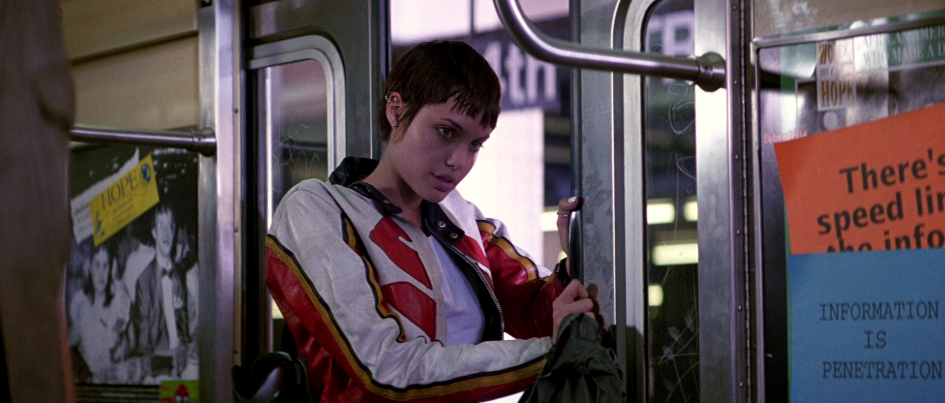 Scene from Hackers (1995) movie: character Kate Libby/Acid Burn played by Angelina Jolie, backpack in hand, standing in closing subway doors, holding them open, in leather Suzuki red and white motorcycle jacket, gazing/beckoning in the character Dade's direction. 'Are you coming?' she asks. Just out of the shot some pieces of orange and blue paper are taped to the subway wall with large printed text. The orange one says There's no speed limit on the information highway. The blue one: Information is penetration. A Cyberdelia sticker can be seen stuck behind the orange paper.