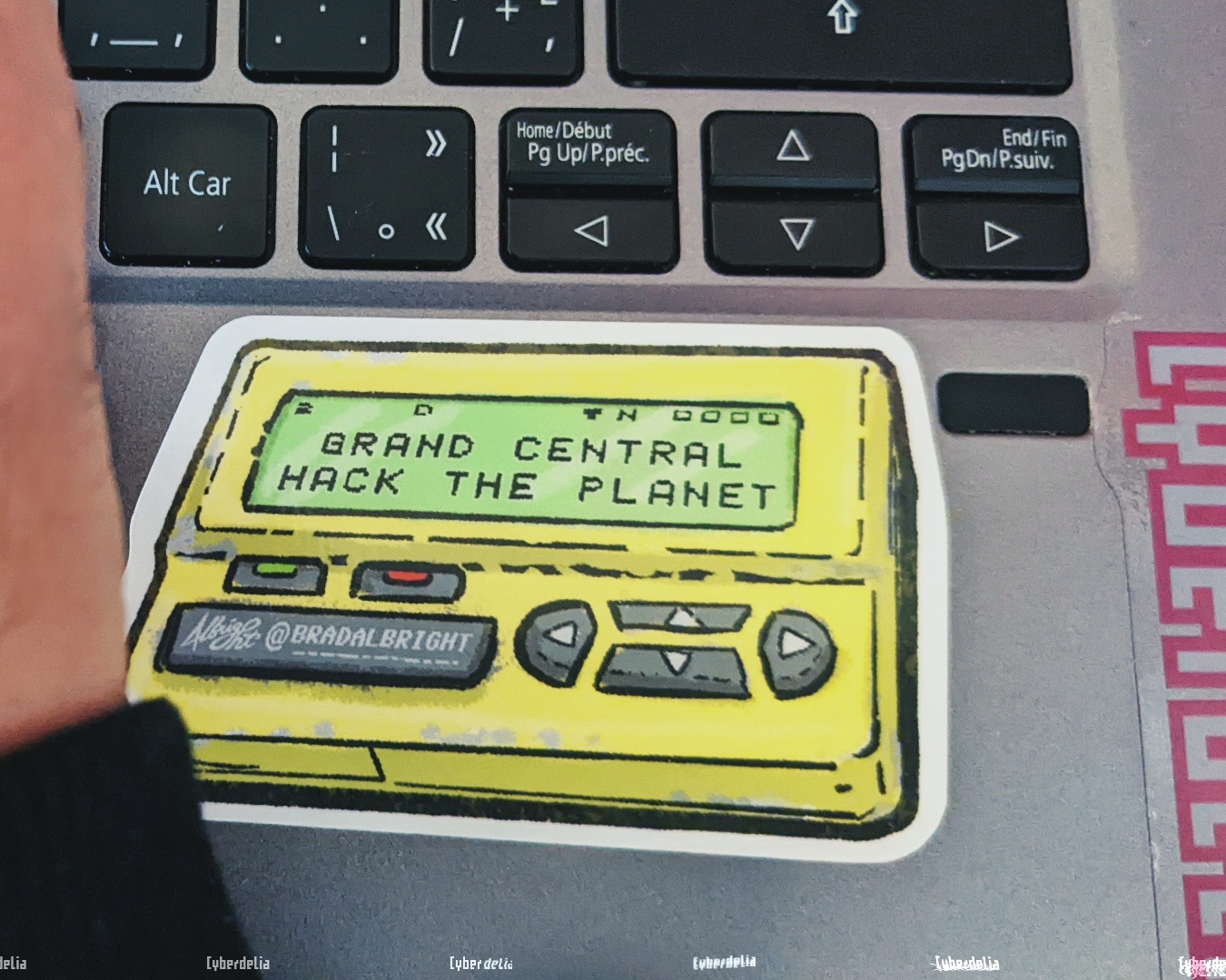 Corner of a laptop keyboard with a sticker on it and edge of a hand typing. Sticker is an illustration of a pager as seen in Hackers (1995) movie, bright yellow with green LCD display and text 'GRAND CENTRAL HACK THE PLANET' on it. Sticker art by Brad Albright. Edge of a Cyberdelia NYC sticker can also be seen on the laptop.
