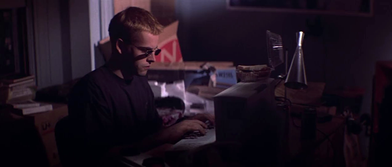 Opening scene of Hackers: Dade Murphy hunched over, typing on a computer terminal in bedroom, sunglasses on, dim lights.