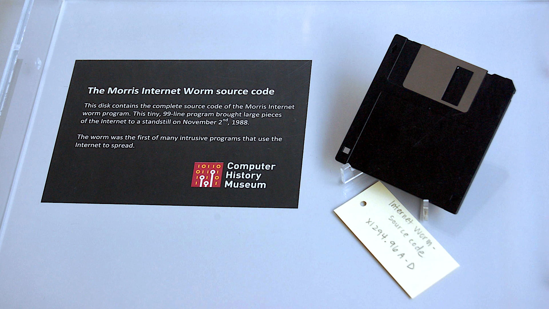 Photo of a display case at the Computer History Museum containing a single black 3.5-inch floppy disk. A yellow paper tag sitting beneath it with handwriting in pencil: Internet Worm - Source Code X1294.96A-D. A black museum description plackard sits to the left: 'The Morris Internet Worm source code - This disk contins the complete source code of the Morris Internet worm program. This tiny, 99-line program brought large pieces of the Internet to a standstill on November 2nd, 1988. The worm was the first of many intrusive programs that use the Internet to spread.' A red square logo is at bottom of plackard with binary ones and zeroes symbols and white text COMPUTER HISTORY MUSEUM. Original photo by the Intel Free Press.
