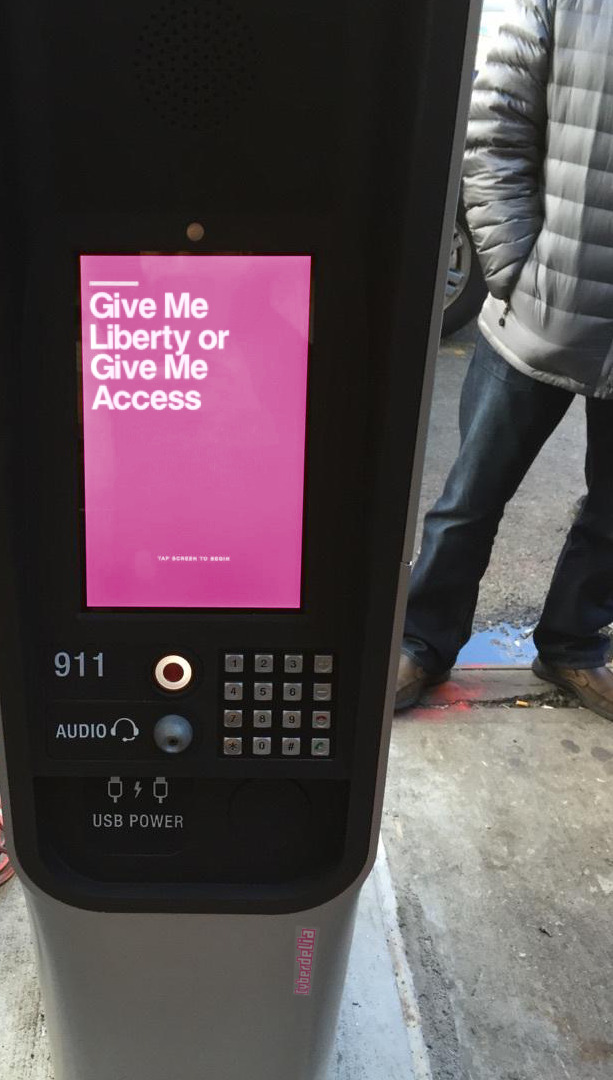 A LinkNYC modern payphone terminal in New York City with features like USB charging port, wifi and digital display screen. This one with a pink image with bold white text 'Give Me Liberty or Give Me Access' as seen in Hackers (1995) movie. A CyberdeliaNYC sticker can also be seen on it.